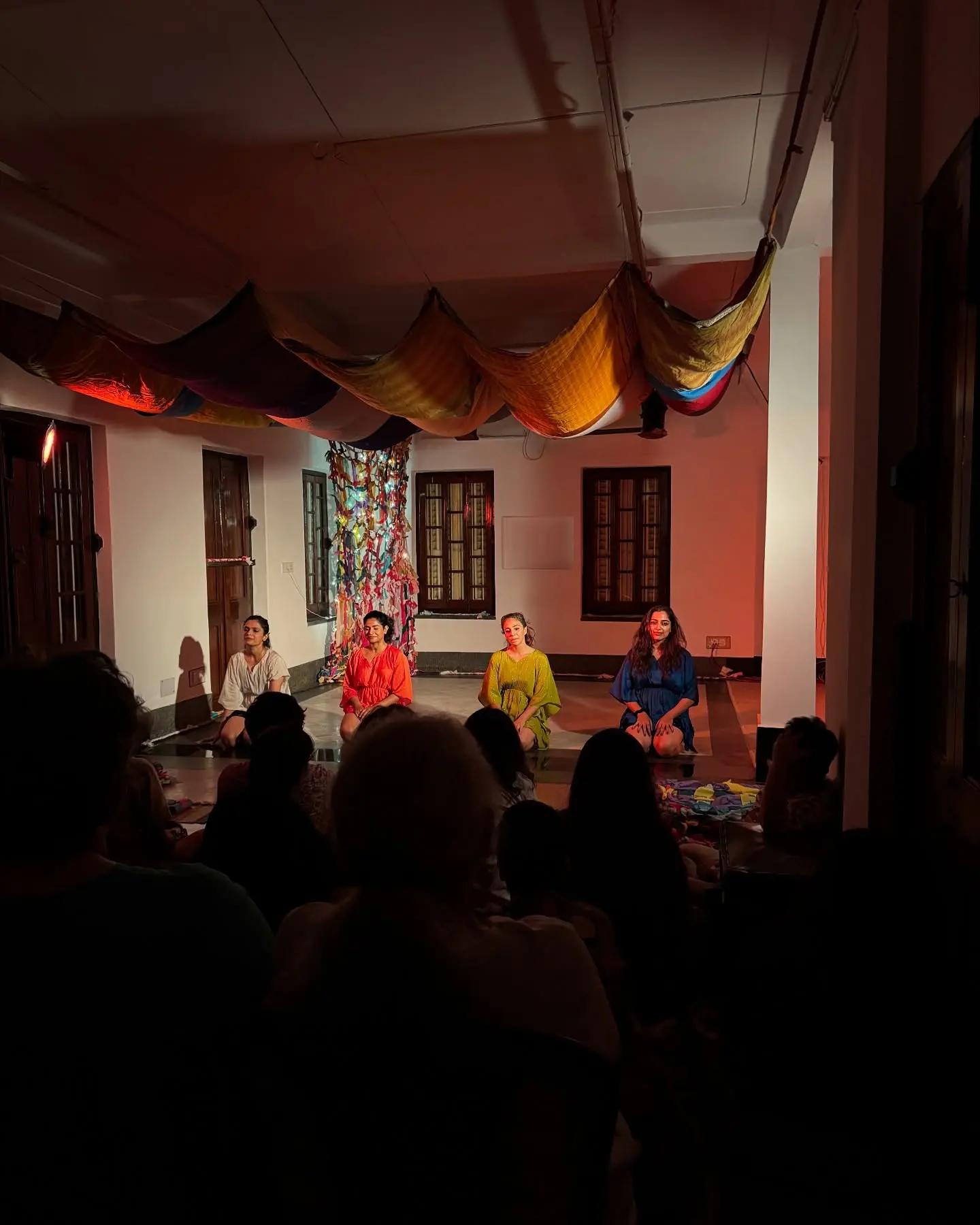 The audience in silhouette on the studio floor, watching a lit stage under draped saris.