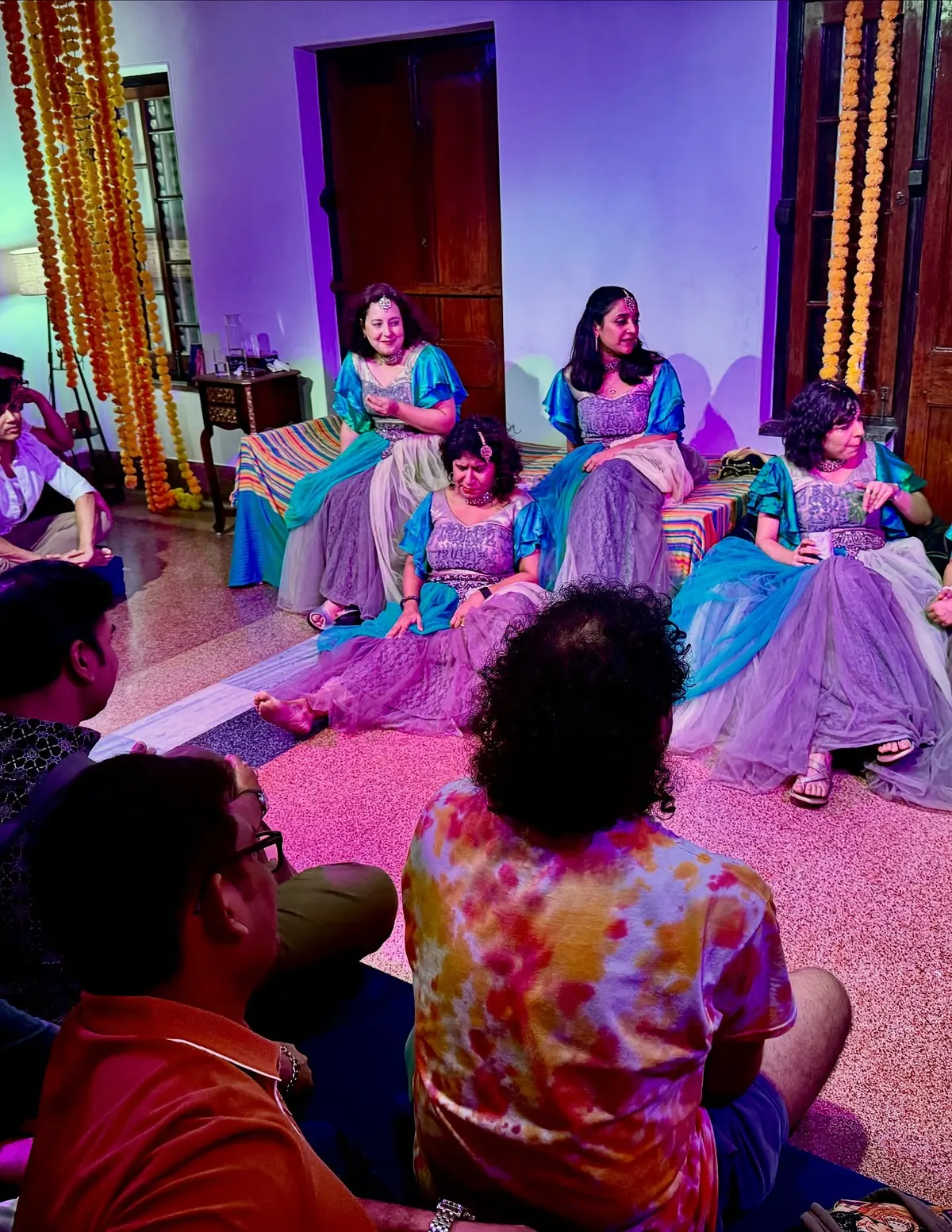 Five women in bridal lehengas under magenta light, the audience on the floor in front of them.